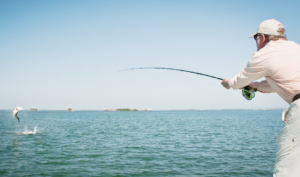 A man with a rod and reel in the foreground while a tarpon leaps out of the water in the background.