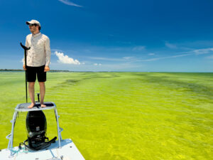 A man stands on a boat that is over a shallow, sandy bottom.